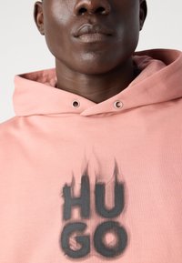 Sudadera de algodón de color rosa con el logo "HUGO" en negro y acentos en forma de llama. Incluye capucha con cordón y ojales de metal.