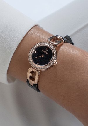 Roségoldene und schwarze Furla-Armbanduhr mit kristallbesetzter Lünette am Handgelenk, schwarzem Lederarmband und schwarzem Zifferblatt mit Logomuster und goldfarbenen Zeigern.