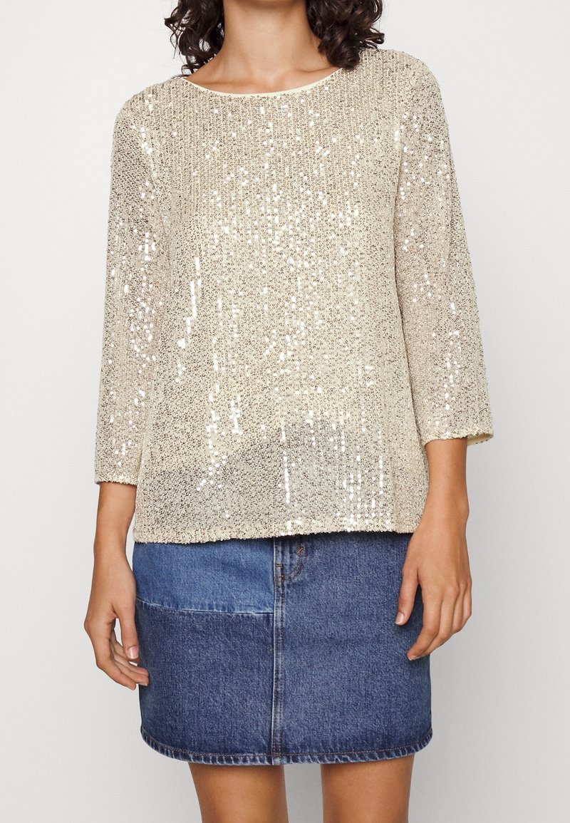 Top beige con paillettes, maniche a trequarti, colletto rotondo, vestibilità ampia, abbinato a una gonna di jeans. Texture scintillante, materiale leggero.
