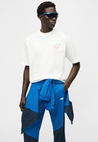 Νεαρός άνδρας που φοράει λευκό T-shirt με το λογότυπο της New Balance, μπλε μπουφάν δεμένο στη μέση, ταιριαστά μπλε και μαύρα παντελόνια και μαύρα γυαλιά ηλίου, κοιτάζοντας πλάγια.