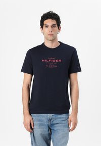 Tamsiai mėlynas medvilninis marškinėliai su apvalia kaklo linija, trumpomis rankovėmis ir raudonu spaudiniu "TOMMY HILFIGER NEW YORK CITY" ant priekio.