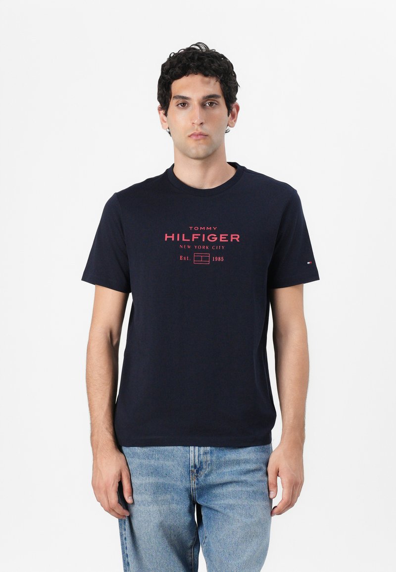 Tamsiai mėlynas medvilninis marškinėliai su apvalia kaklo linija, trumpomis rankovėmis ir raudonu spaudiniu "TOMMY HILFIGER NEW YORK CITY" ant priekio.