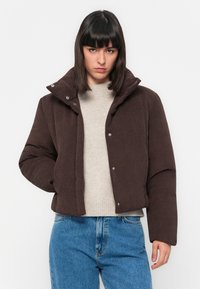 Donna che indossa un giubbotto imbottito marrone scuro sopra un maglione beige e jeans blu, in piedi di fronte a uno sfondo bianco semplice.