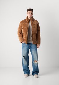 Braune cordartige Pufferjacke mit hohem Kragen und Reißverschluss, getragen über einem beigen Pullover und verwaschenen hellblauen Jeans. Helle Schuhe.