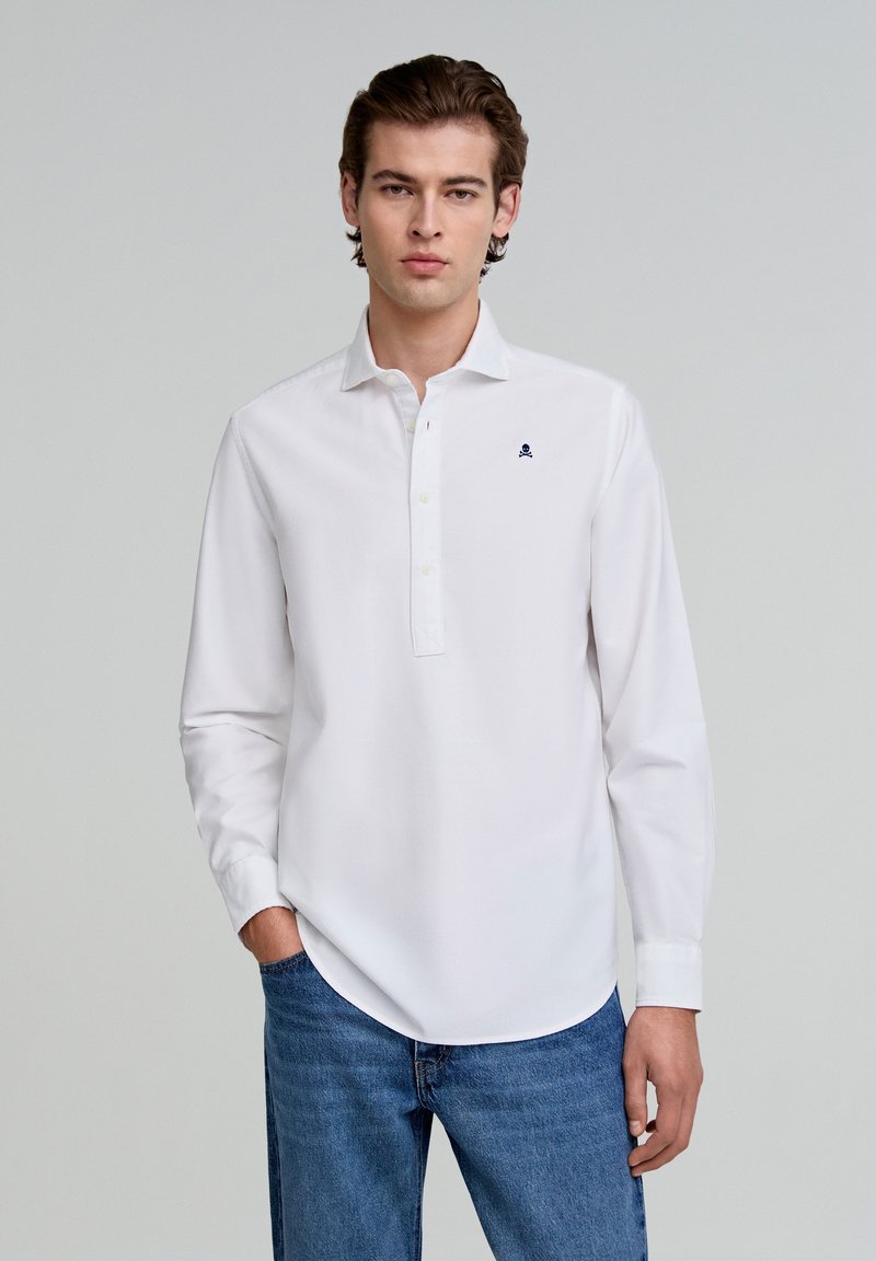 Camisa blanca de manga larga con tapeta de botones, que presenta un pequeño logotipo azul marino en el pecho. Combinada con jeans azules, que muestran una textura suave.