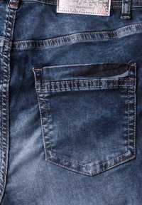 Jean en denim bleu foncé avec une finition texturée, comportant une poche arrière et des coutures contrastées. Étiquette de marque en argent à la taille.