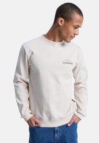 Sudadera beige clara con mangas largas y puños acanalados. Presenta un logo sutil en el pecho y un acento en la costura lateral. Textura suave.