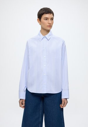 Person with short hair wearing a light blue and white striped button-up shirt and wide-leg dark blue jeans, standing against a plain background.