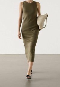 Robe midi sans manches vert olive avec une silhouette ajustée, associée à un sac beige texturé et des sandales noires à enfiler. Fini en tissu lisse.