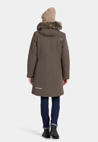 Person, der einen braunen Wintermantel mit Pelzhut, eine beige Strickmütze, dunkle Jeans und schwarze Stiefel trägt, steht mit dem Rücken zu einem weißen Hintergrund.