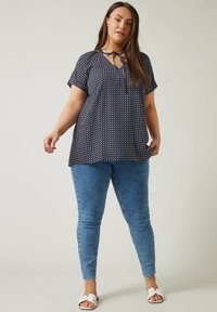 Dunkelblaue Bluse mit weißen Punkten, kurzen Ärmeln und einem Schlüsselaugen-Halsband, kombiniert mit hellblauen Skinny-Jeans und weißen Sandalen.