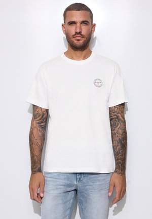 Hombre con cabello corto y tatuajes en ambos brazos, vistiendo una camiseta blanca y vaqueros azul claro, de pie contra un fondo blanco.