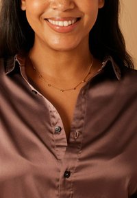 Gold necklace with small heart and round accents, worn over a smooth, beige-brown button-up shirt with a classic collar.