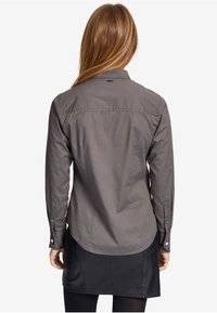 Grå langærmet skjorte med knapper og buet kant, med metalhardware og kontrasterende knapdetaljer på manchetterne. Blød tekstur.