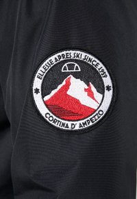 Svart stoff har et sirkulært brodert merke med et rødt og hvitt fjelldesign, "Cortina D'Ampezzo," og "Ellesse Apres Ski Since 1959."