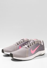 Scarpe sportive con tomaia in rete di colore grigio chiaro, caratterizzate da un audace logo Nike swoosh in rosa. Suola bianca, dettagli di design testurizzati e accenti a contrasto.