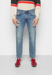 Persona que lleva puesto unos vaqueros claros desgastados, zapatillas rojas y blancas, y una camiseta de manga larga con rayas beige, negras y rojas.