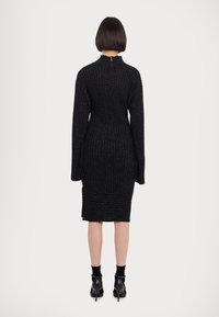 Femme aux cheveux courts et foncés portant une robe midi en maille côtelée noire, des chaussettes noires et des chaussures à talons noirs, debout devant un fond uni.