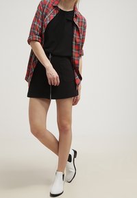 Camicia a quadri rossi e neri sopra un vestito nero senza maniche con dettaglio a fiocco; abbinata a stivaletti neri e bianchi alla caviglia.