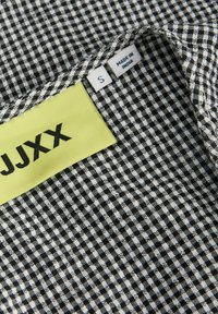 Tissu à carreaux noir et blanc montrant une étiquette de marque JJXX jaune et deux étiquettes blanches marquées "S" et "Made in India" sur le col du vêtement.