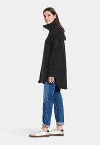 Sort fleece-pullover med høj krave, sidepaneler og et præget logo på ærmet. Parret med blå jeans med opfoldede ærmer og hvide sneakers.