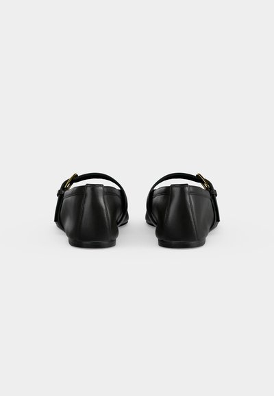 Paire de chaussures plates noires avec brides à la cheville et boucles dorées, vues de dos sur un fond uni clair.