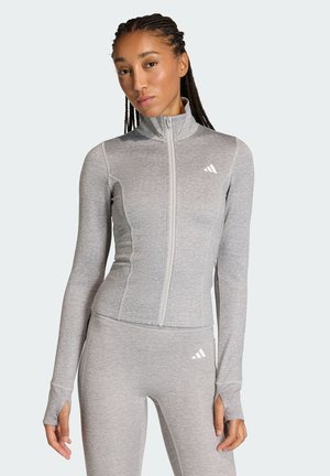 Frau mit geflochtenem Haar, die eine hellgraue, taillierte Reißverschlussjacke und passende Hosen mit Daumenlöchern und Adidas-Logos trägt.