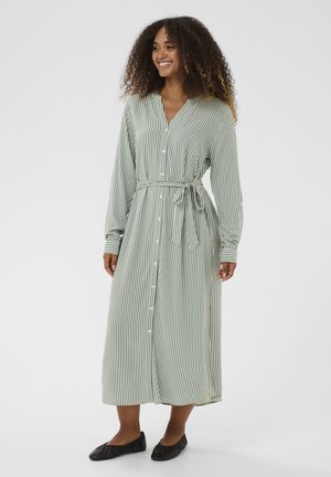 Grün-weiß gestreiftes Hemdblusenkleid mit Bindegürtel in der Taille, langen Ärmeln und midikurzer Länge. Weicher Stoff mit lockerem Schnitt. Schwarze flache Schuhe.