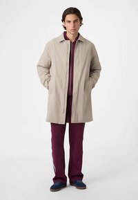 Beige letvægtsfrakke med knaplukning og sidelommer, over et burgunder outfit med hvide sidelister, sat sammen med blå og brune sko.