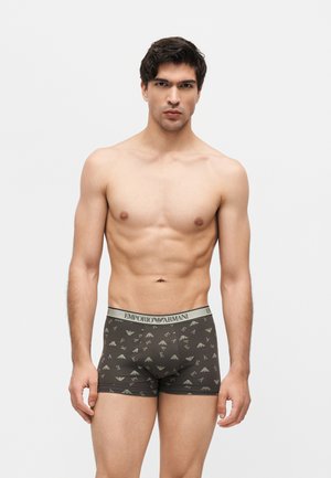 Młody muskularny mężczyzna stojący, ubrany w ciemne, wzorzyste bokserki Emporio Armani, na jednolitym jasnoszarym tle.