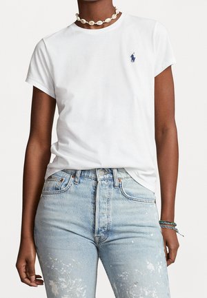 Personne portant un t-shirt blanc à manches courtes avec un logo bleu, un jean bleu clair éclaboussé de peinture, un collier de coquillages et des bracelets au poignet gauche.