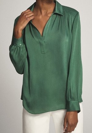 Blusa verde a maniche lunghe in tessuto liscio; presenta un colletto con bottoni, dettagli plissettati sulle spalle e polsini arricciati.
