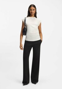 Lys beige tætsiddende top med kasketærmer, kombineret med højtaljede, sorte bukser med brede ben. En lille sort skuldertaske bæres på den ene side.