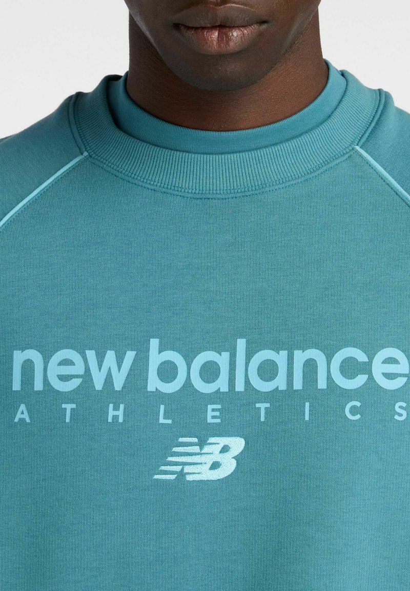 Primo piano di una persona che indossa una felpa verde acqua con la scritta "new balance ATHLETICS" e il logo New Balance sul davanti.