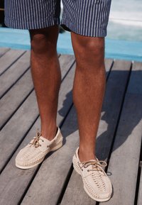 Mocassins tissés beige avec une surface texturée, des détails tressés, et des lacets clairs, portés avec un short à rayures bleu marine sur une terrasse en bois.