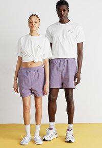 Weiße kurzärmelige T-Shirts mit kleiner Stickerei, kombiniert mit lila Shorts. Das männliche Modell trägt weiße und lila Sneakers, während das weibliche Modell weiße Schuhe trägt.