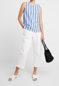 Top senza maniche a righe blu e bianche abbinato a pantaloni bianchi a gamba larga e sandali bianchi con punta aperta. Una piccola borsa a tracolla nera intrecciata completa il look.