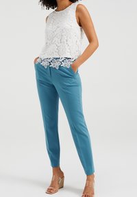 Valkoinen pitsinen hihaton toppi, jossa on kukkakuvio ja sahalaitainen helma, yhdistettynä vaaleansinisiin muotitakkeihin ja läpinäkyviin korkokenkiin.