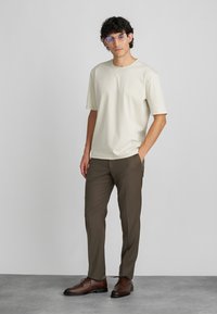 Ανοιχτό μπεζ κοντομάνικη μπλούζα συνδυασμένη με σκούρο καφέ παντελόνι. Το μοντέλο φορά καφέ παπούτσια και γυαλιά με μπλε απόχρωση, στέκεται σε γκρι δάπεδο.