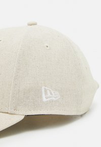 Casquette beige en tissu texturé avec une visière courbée et un logo blanc brodé sur le côté. Comprend des œillets pour la ventilation.
