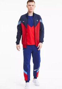 Puma sportieve trainingsoutfit met een marineblauwe jas met rode en blauwe accenten, gecombineerd met een rood shirt en marineblauwe broek met kleurblokelementen.