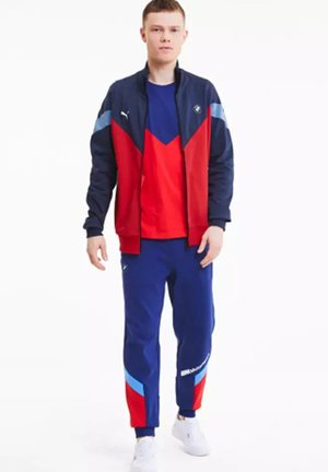 Chándal deportivo de Puma que incluye una chaqueta azul marino con acentos en rojo y azul, combinada con una camiseta roja y pantalones azul marino con detalles en bloques de color.