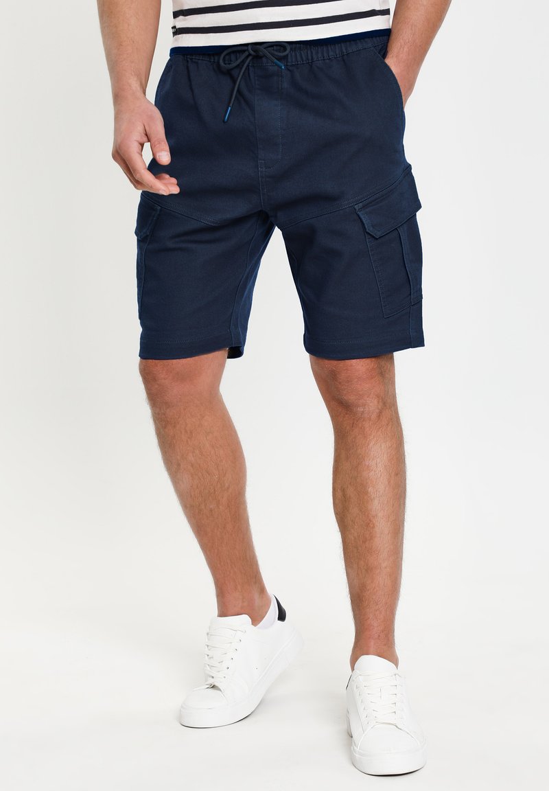 Mand iført navy blå cargo shorts og hvide sneakers, står med hænderne i lommen foran en ensfarvet baggrund.