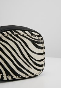 Černobílá kabelka se vzorem zebry a texturou umělé kožešiny, s stříbrnými cvoky podél okraje a zaobleným čtvercovým tvarem.