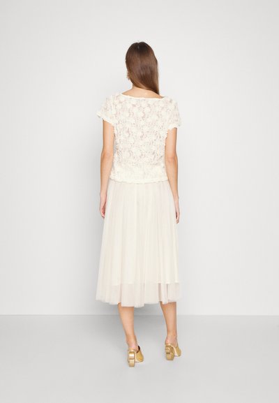 Top din dantelă crem cu mâneci scurte, asortat cu o fustă fluidă din tul transparent, creând un aspect stratificat cu texturi delicate.