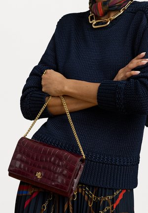 Cilvēks valkā tumši zilu adītu džemperi un plektu svārkus ar ķēdes rakstu, turot bordo krāsas krokodila ādas roku maisiņu ar zelta ķēdes siksniņu un RL logo.