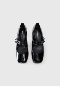 Ein Paar schwarze Lackleder-Mary-Jane-Schuhe mit quadratischen Schuhspitzen und Schnallenriemen, Marke Michael Kors, von oben auf einem weißen Hintergrund betrachtet.