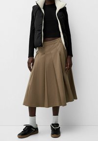 Black zip-up puffer vest over a black long-sleeve top, paired with a tan pleated midi skirt and black sneakers with white socks.