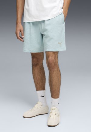 Uomo che indossa pantaloncini celeste chiaro con trama, calzini Puma bianchi e sneakers Puma bianche, in piedi con una mano in tasca.