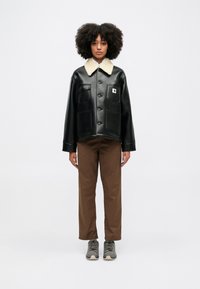 Czarna kurtka z sztucznej skóry z beżowym futrzanym kołnierzem, dużymi kieszeniami z przodu i zapięciem na guziki, w połączeniu z brązowymi spodniami i szarymi butami.